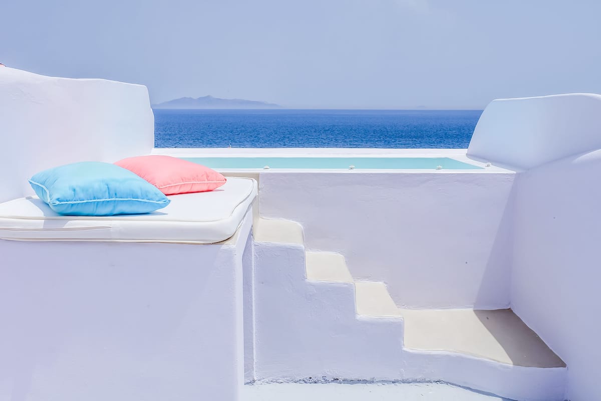 A white outdoor seating area with steps leading to a pool, overlooking the sea.