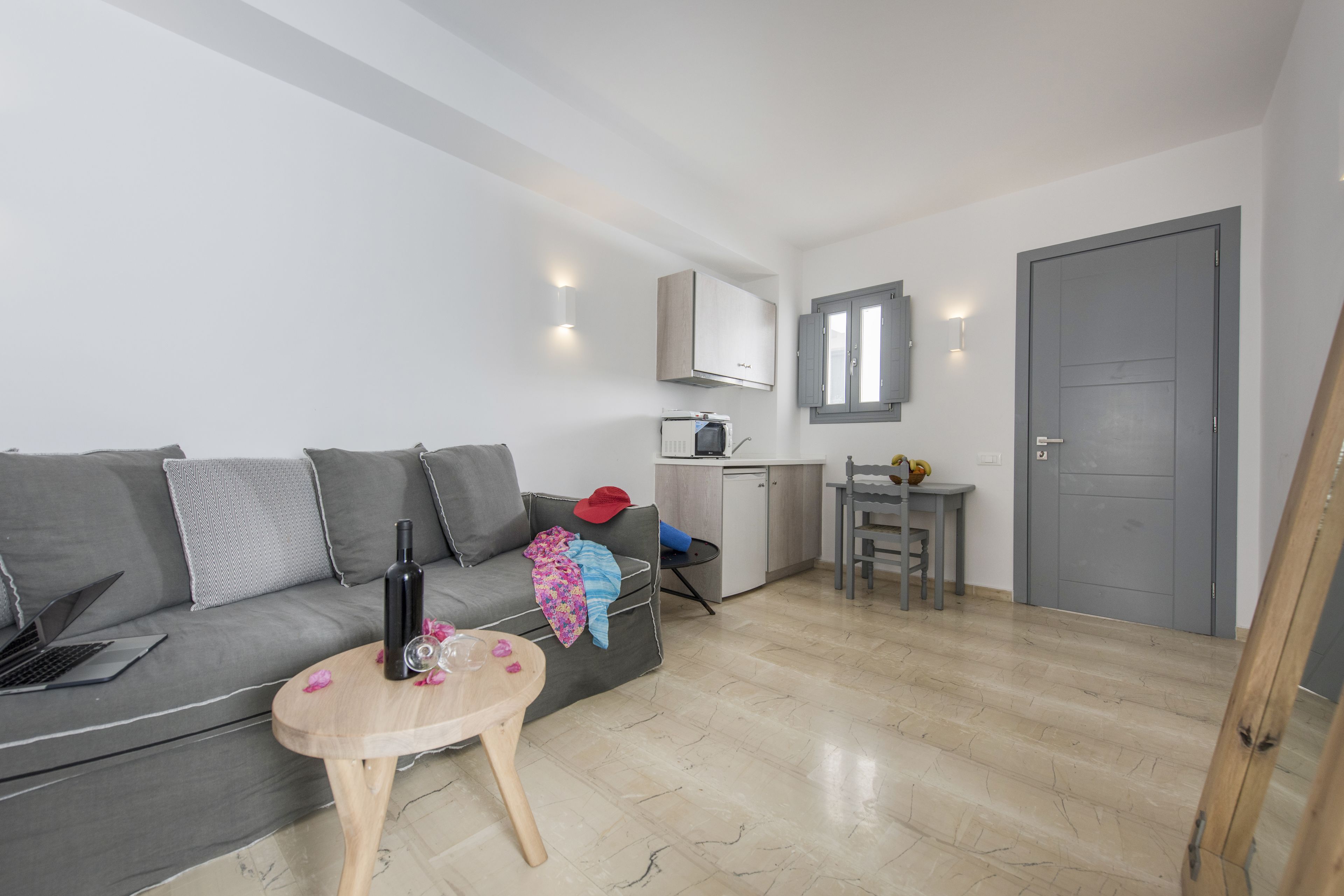 A modern living room with a grey sofa, a wooden coffee table, and a laptop on the sofa. There is a small kitchen area with a microwave and a cabinet.