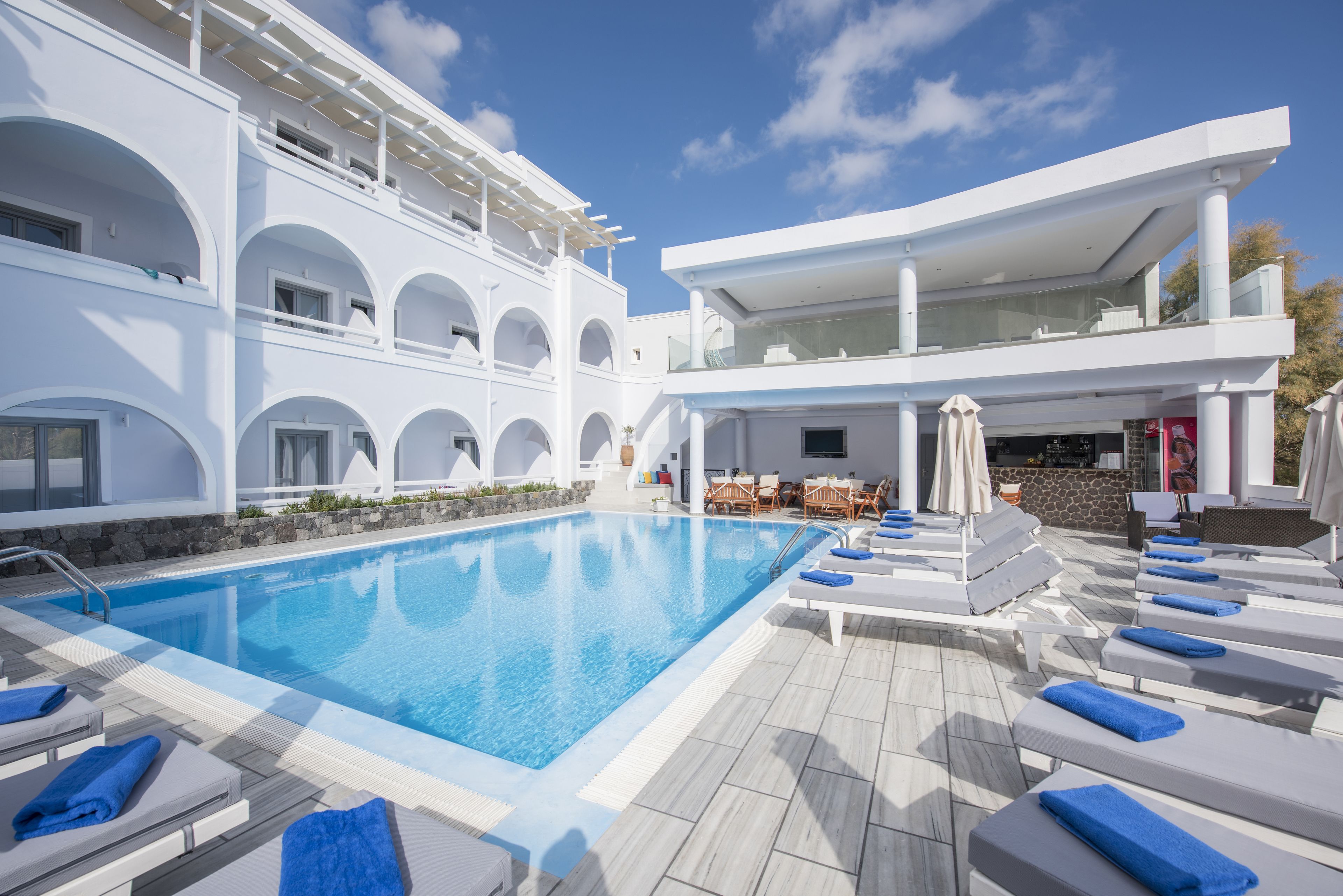 Un área de piscina con sillas de descanso y sombrillas, una piscina azul cristalina y un edificio blanco con varios balcones.