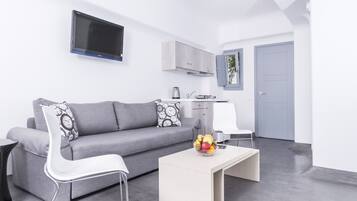 A modern living room with a grey sofa, white armchair, and a coffee table with fruit. A TV is mounted on the wall, and a kitchen area is visible in the background.