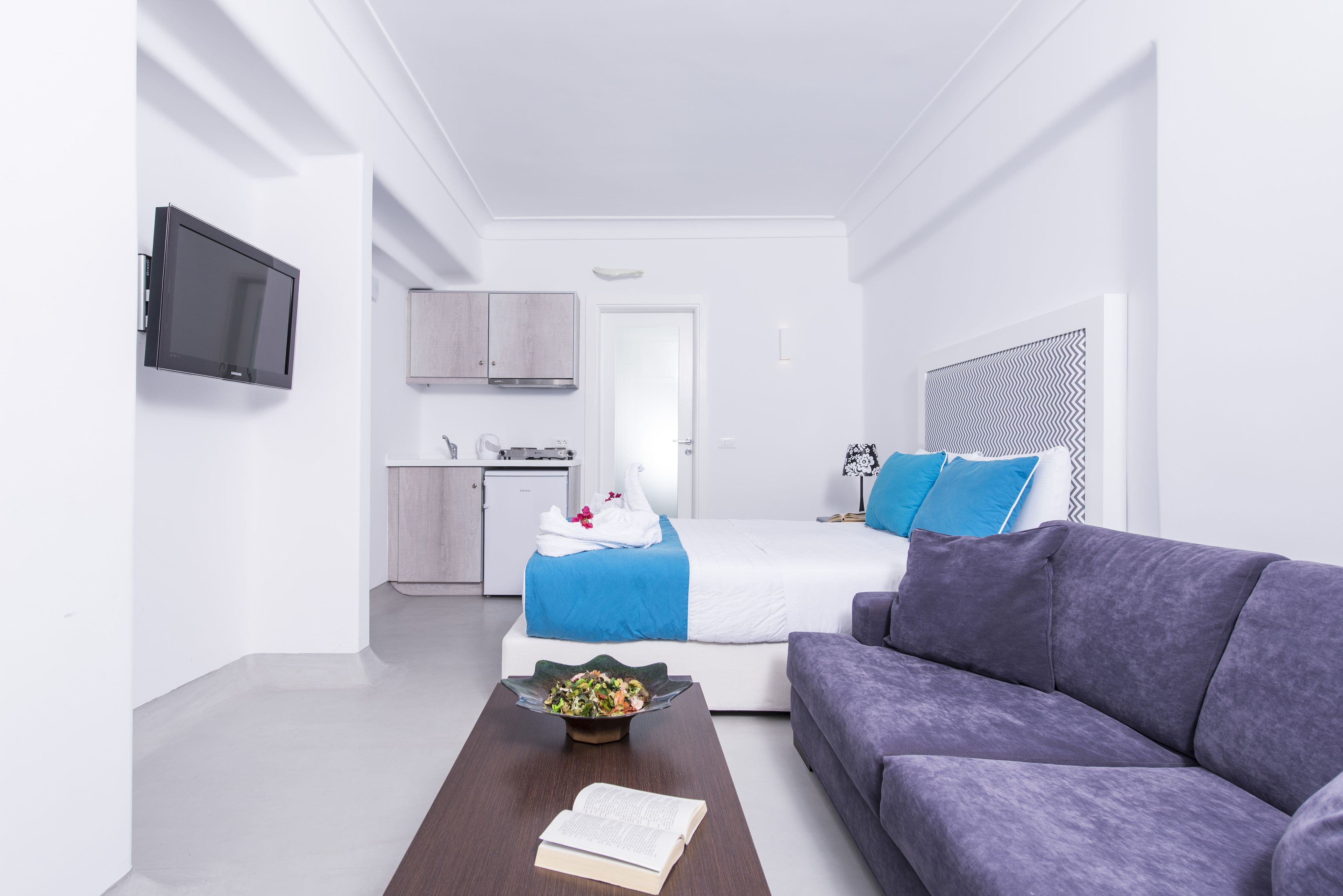 A modern hotel room with a bed, a sofa, a dining table with a book and a bowl of fruit, a TV, a kitchenette, and a patterned headboard.