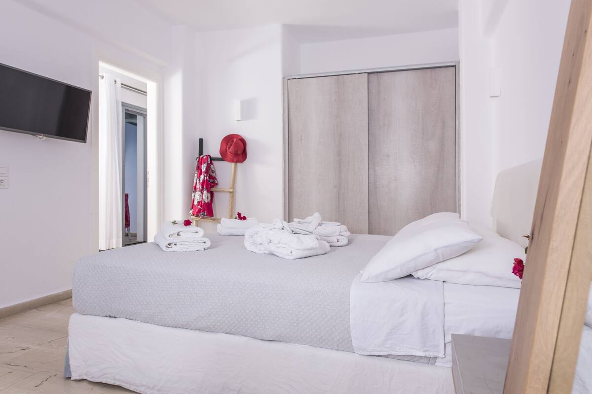 A neatly made bed with white linens, a television mounted on the wall, and a wardrobe with a light wood finish.