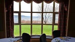 Una habitación con dos mesas de comedor dispuestas para dos personas, un ventanal con cortinas y vistas a un campo cubierto de césped y árboles.