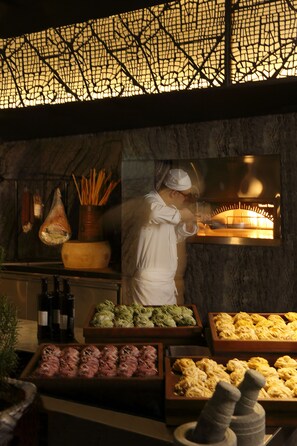 A chef in a kitchen with a wood-fired oven, various food items, and kitchen utensils. - Hyatt Regency Gurgaon (Gurugram)