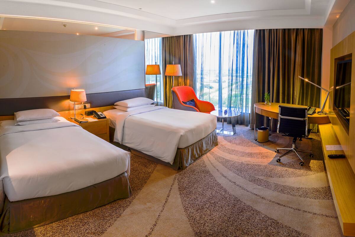 Habitación de hotel con dos camas, un escritorio, una silla, un televisor y un ventanal con cortinas.
