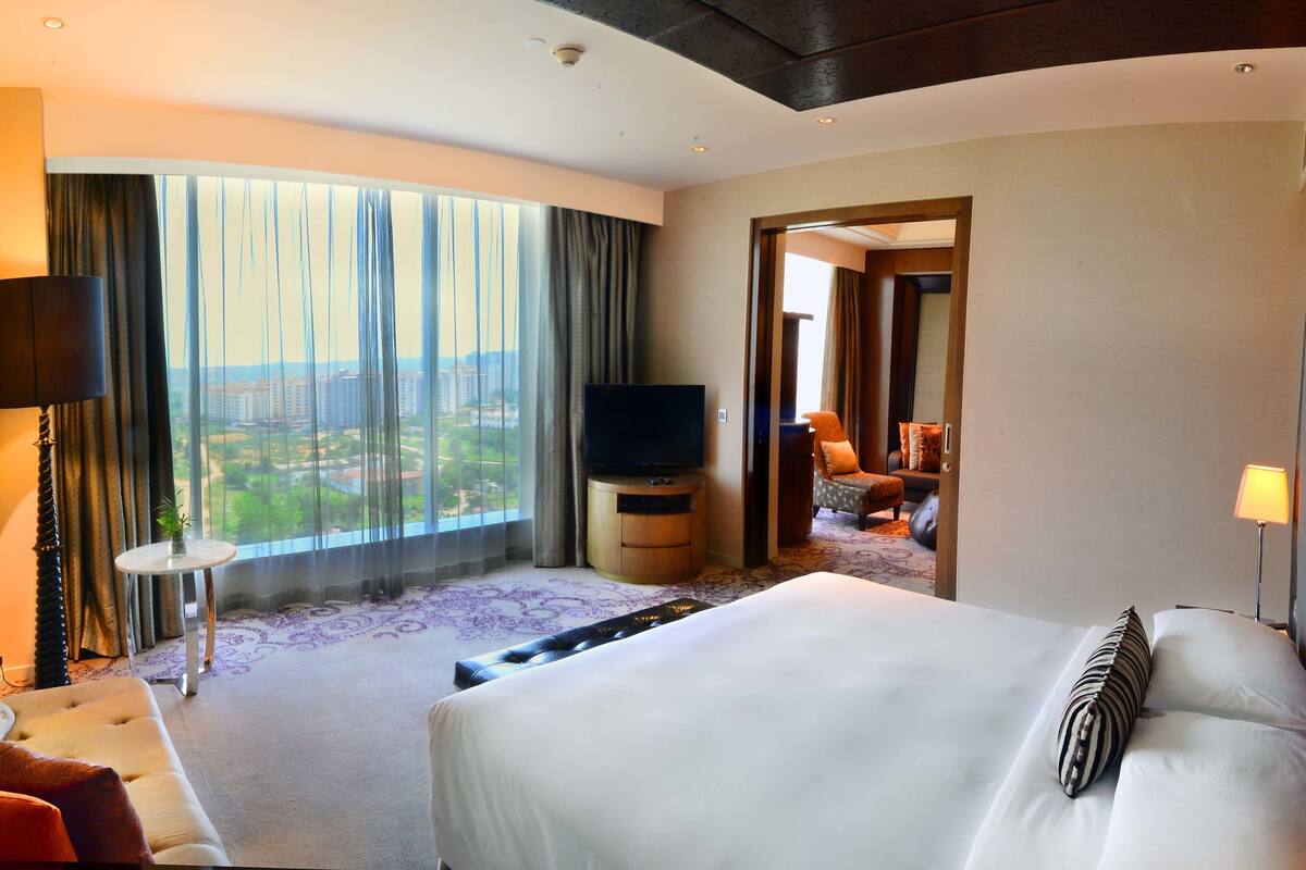Habitación de hotel con una cama grande, televisión, vistas a la ciudad y una mesita con lámpara.