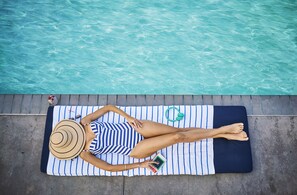 Una persona recostada sobre una toalla a rayas junto a una piscina, con un libro y una botella de agua verde cerca.
