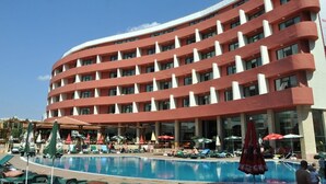 A hotel with a curved facade, a swimming pool, and outdoor seating with umbrellas. - Mena Palace (Sunny Beach)