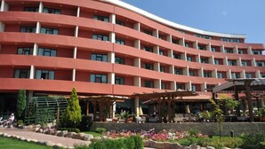 A multi-story hotel with a curved facade, outdoor seating areas, and a garden with flowers and shrubs. - Mena Palace (Sunny Beach)