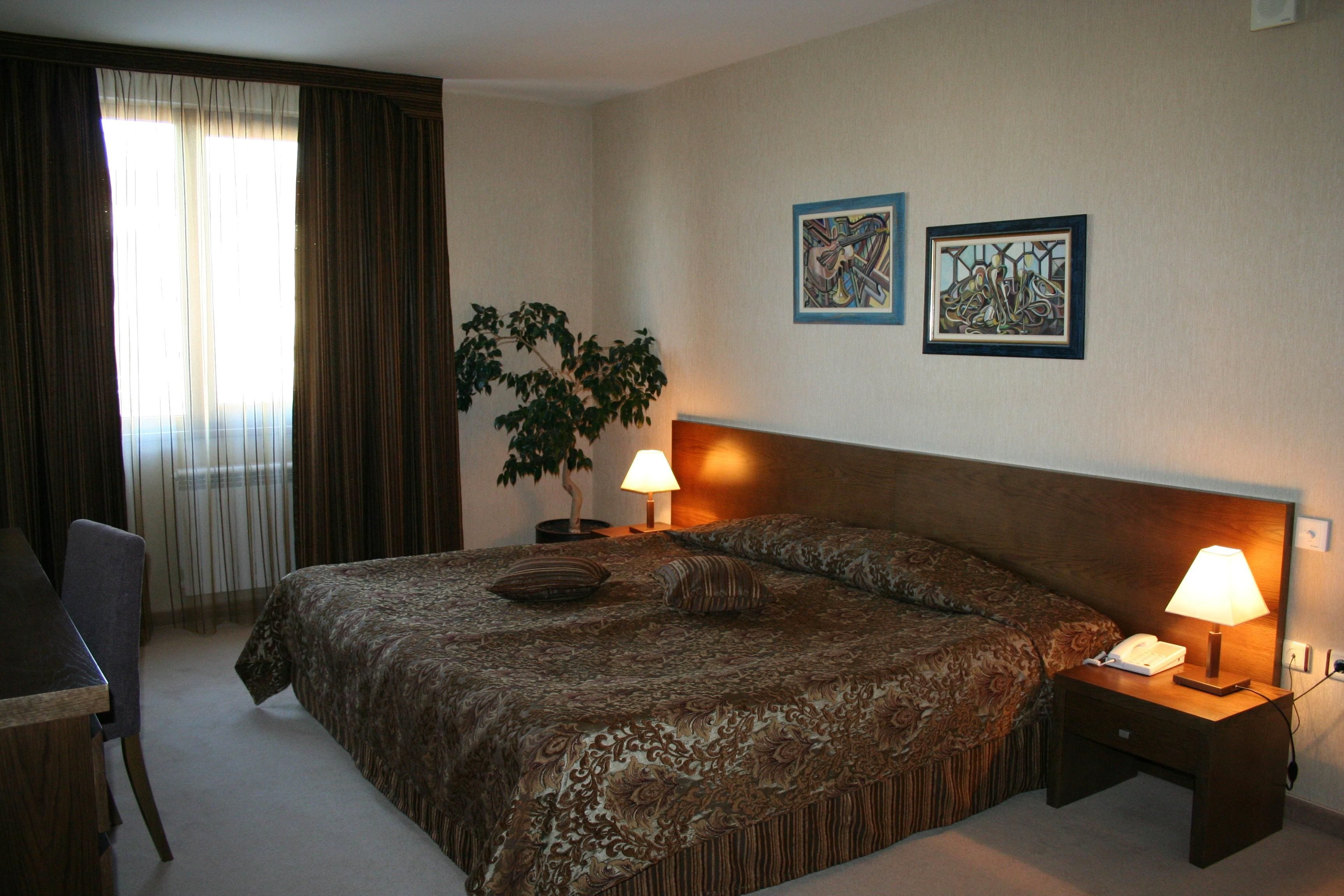 A hotel room with a large bed, two bedside lamps, a potted plant, and two framed artworks on the wall.