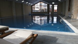 En inomhuspool med klart vatten, en solstol i trä och utsikt över byggnader genom stora fönster.