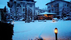 En snötäckt innergård med en byggnad i bakgrunden, en liten konstruktion med ett snötäckt tak och en upplyst lyktstolpe.