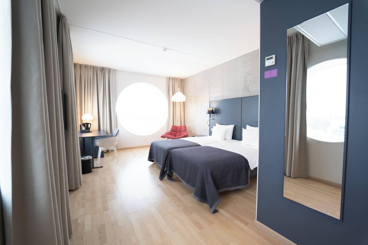 Ein modernes Hotelzimmer mit einem großen Bett, einem Schreibtisch mit Stuhl, einem runden Fenster und einem Spiegel an der Wand.