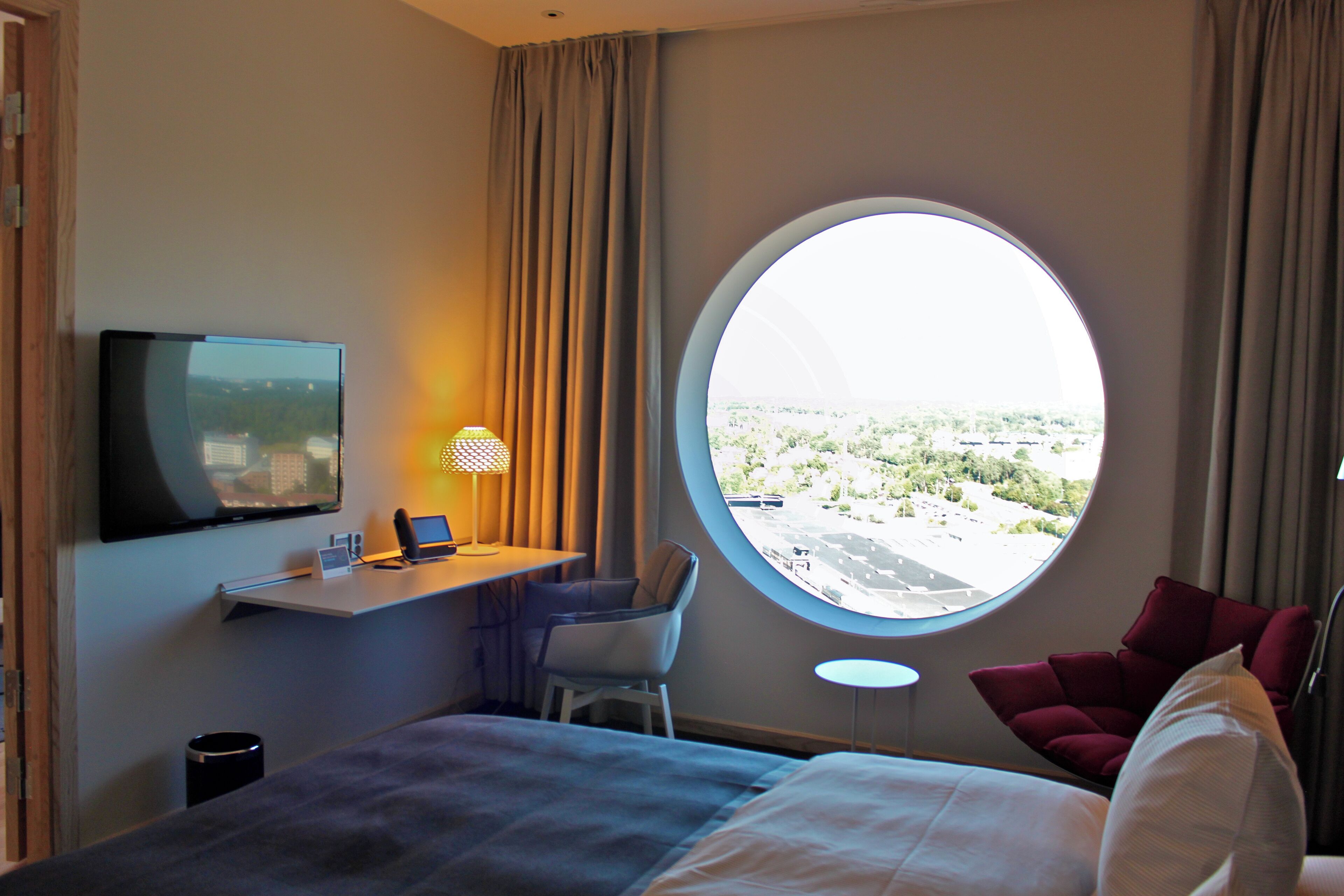 Una habitación de hotel con una cama, un escritorio con una computadora portátil, un televisor, una ventana redonda con vistas, una silla y un sillón rojo.