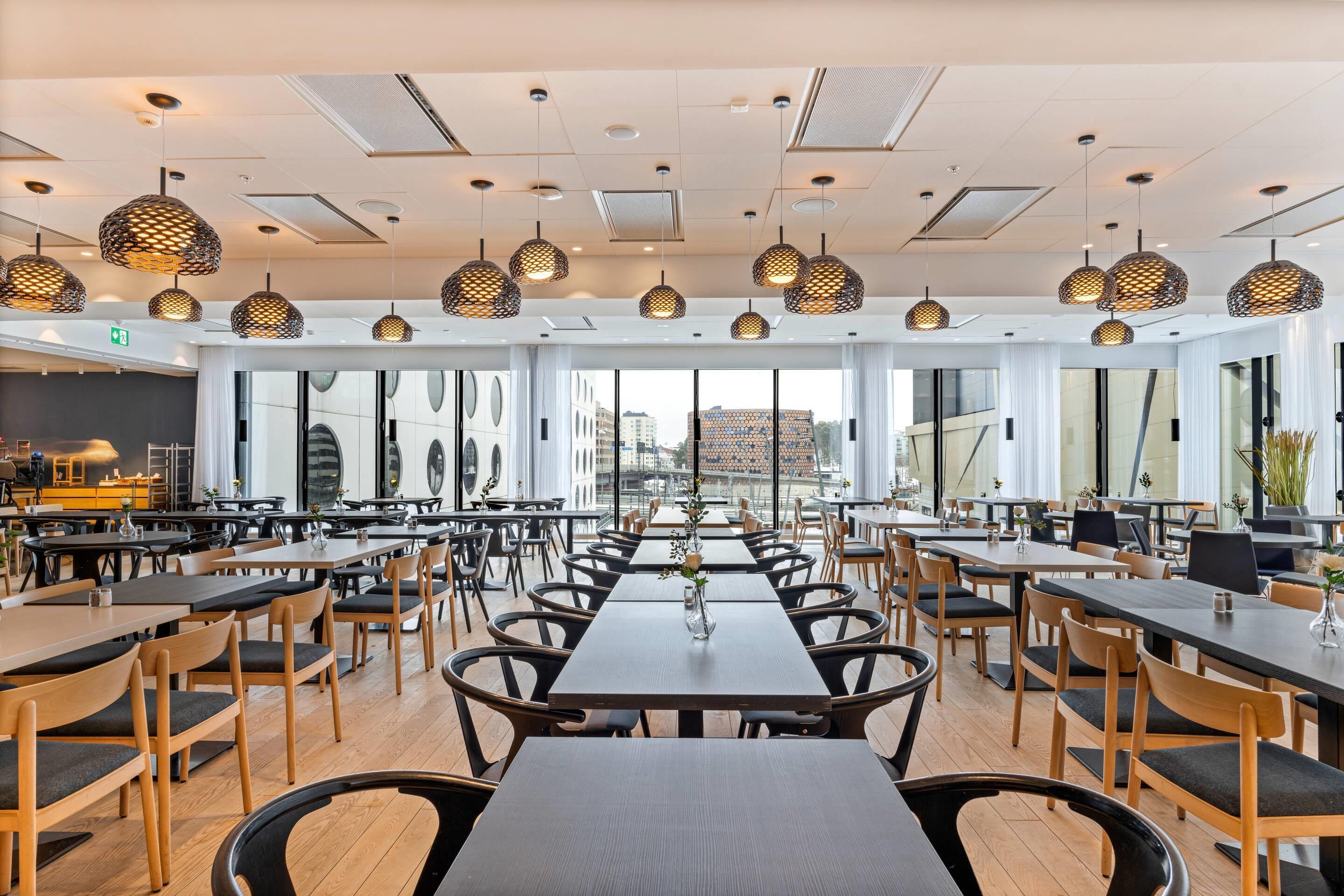 A spacious dining area with multiple tables and chairs, large windows offering views of the city, and modern pendant lighting.