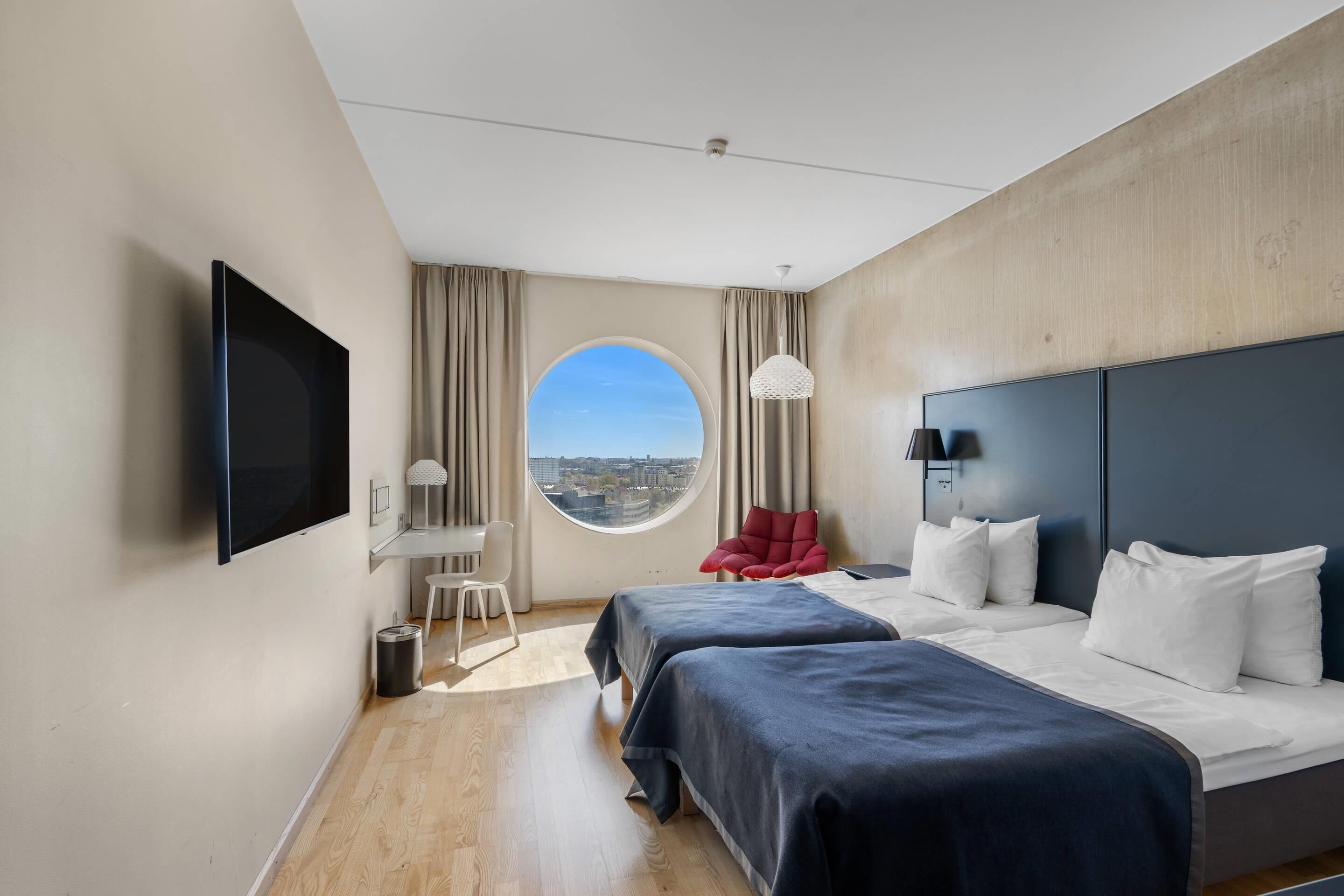 A modern hotel room with a large bed, a TV, a round window, and a view of the city.