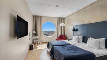 A modern hotel room with a large bed, a TV, a round window, and a view of the city.