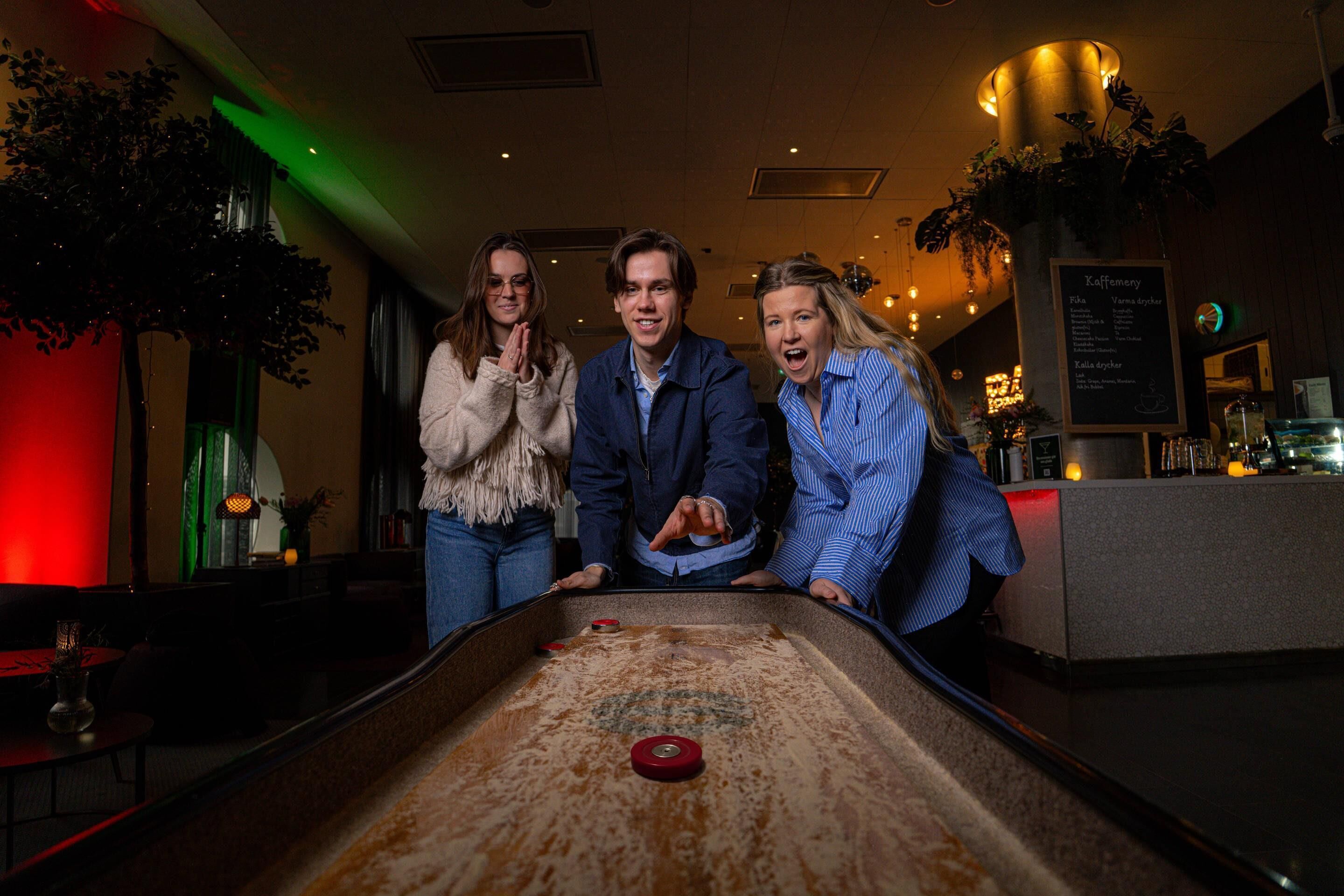 Tres personas jugando a la petanca en una habitación con poca luz, con una zona de bar al fondo.