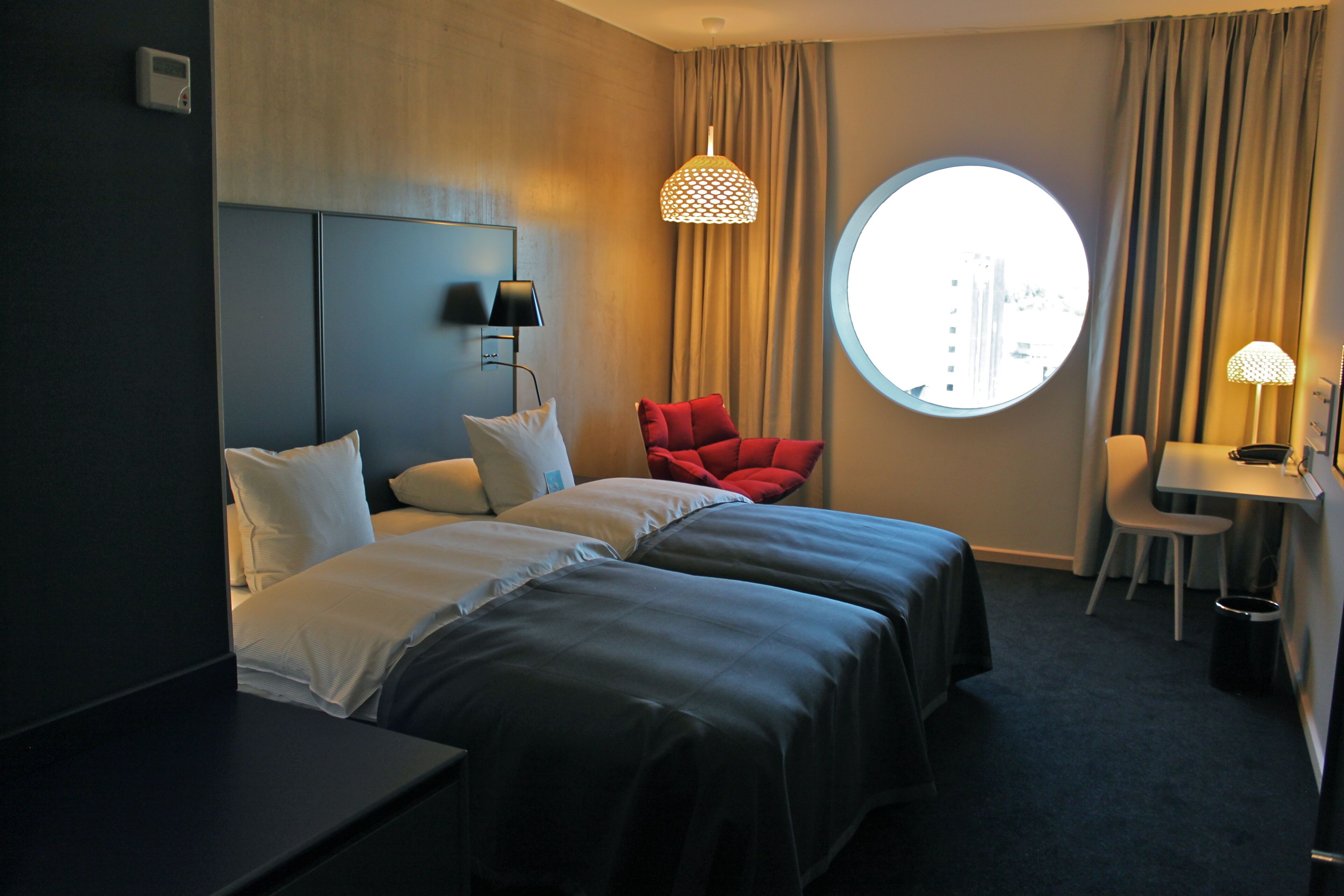 Una habitación de hotel con dos camas, una silla roja, un escritorio, una lámpara y un ventanal redondo.