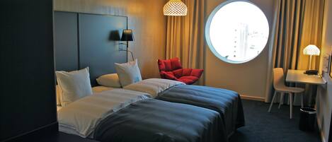 Una habitación de hotel con dos camas, una silla roja, un escritorio, una lámpara y un ventanal redondo.