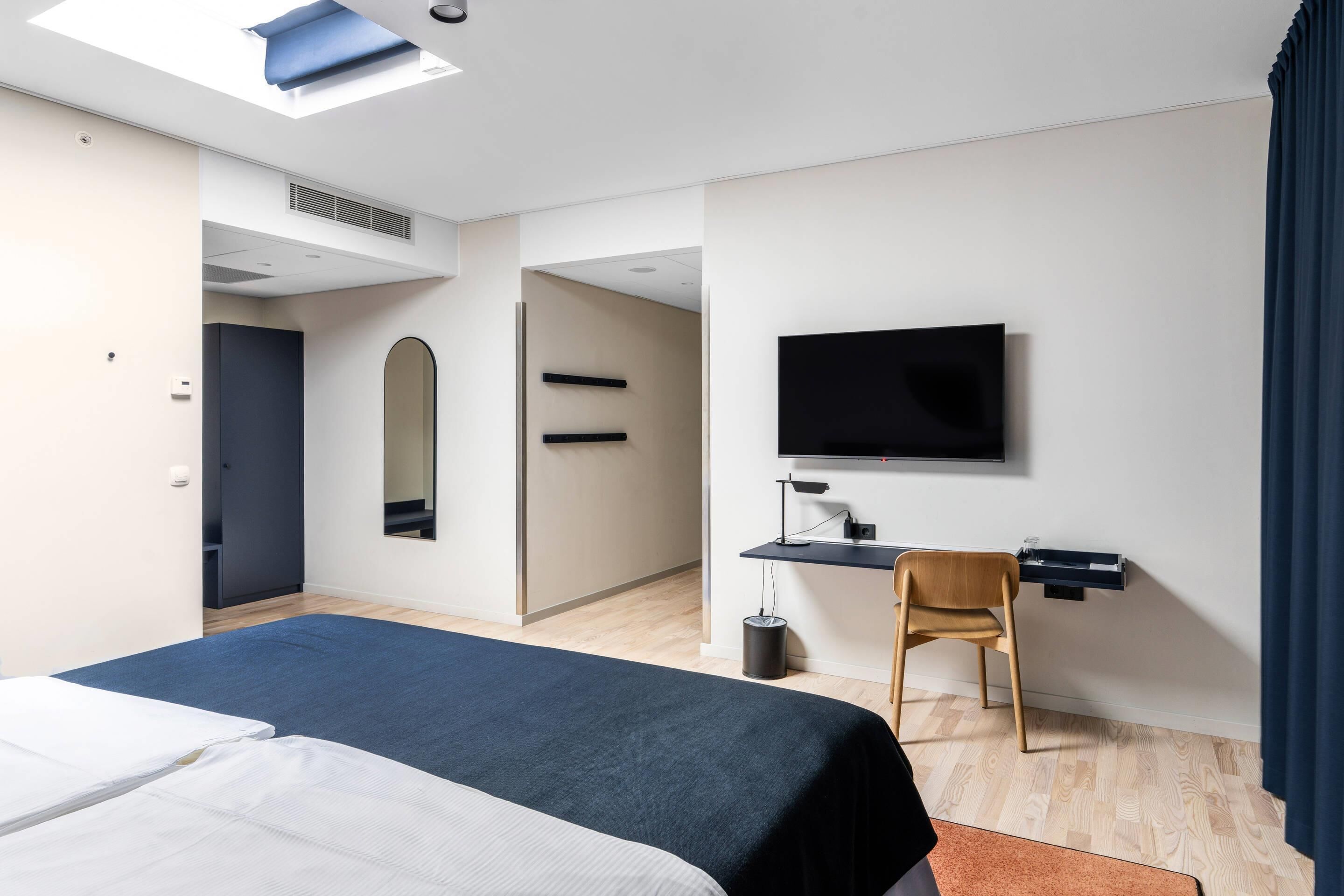 Et moderne hotelværelse med en seng, et skrivebord, en stol, et fjernsyn og et stort spejl.