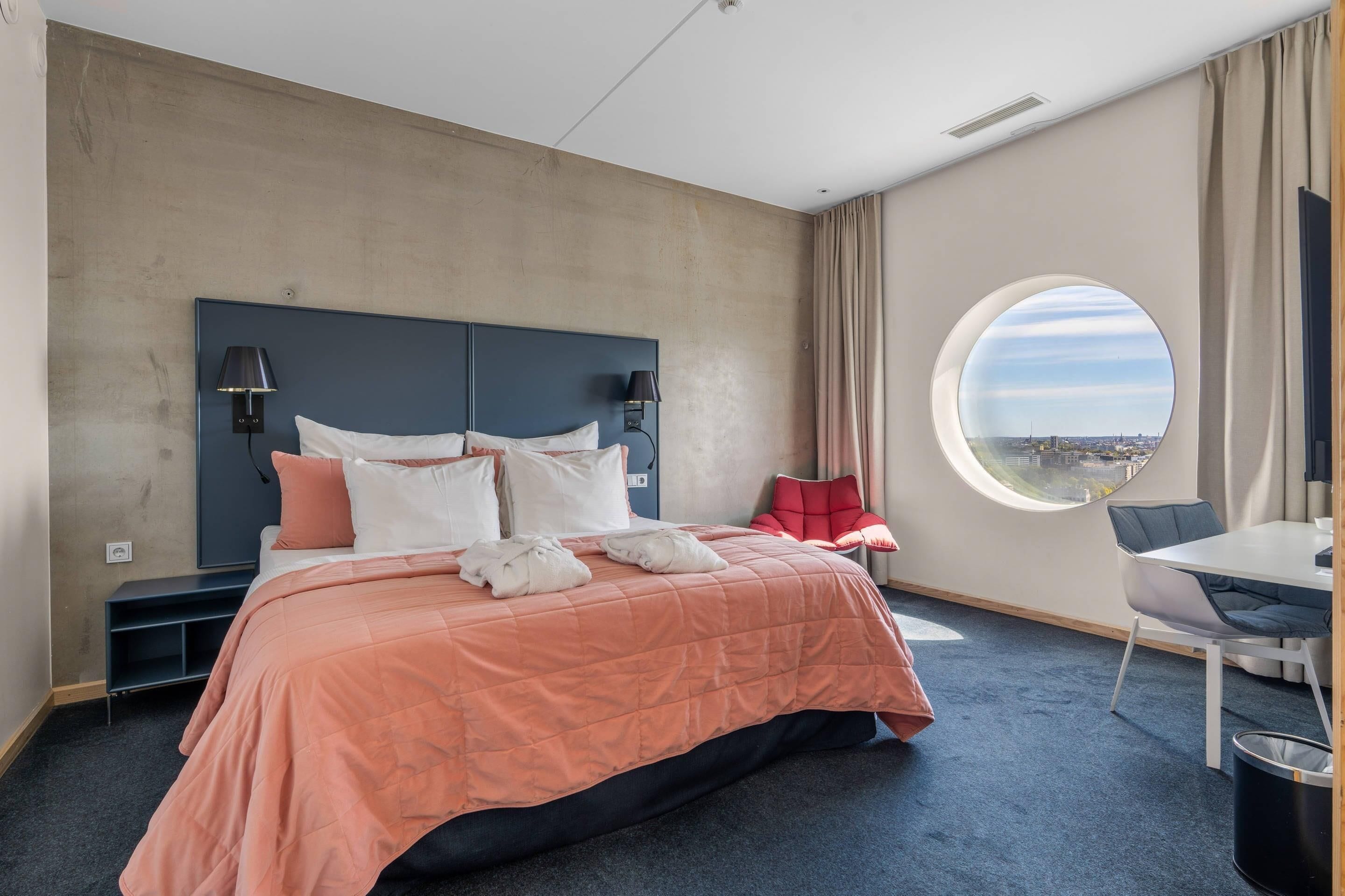 A hotel room with a bed, a round window with a view, a desk with a chair, and a small table.
