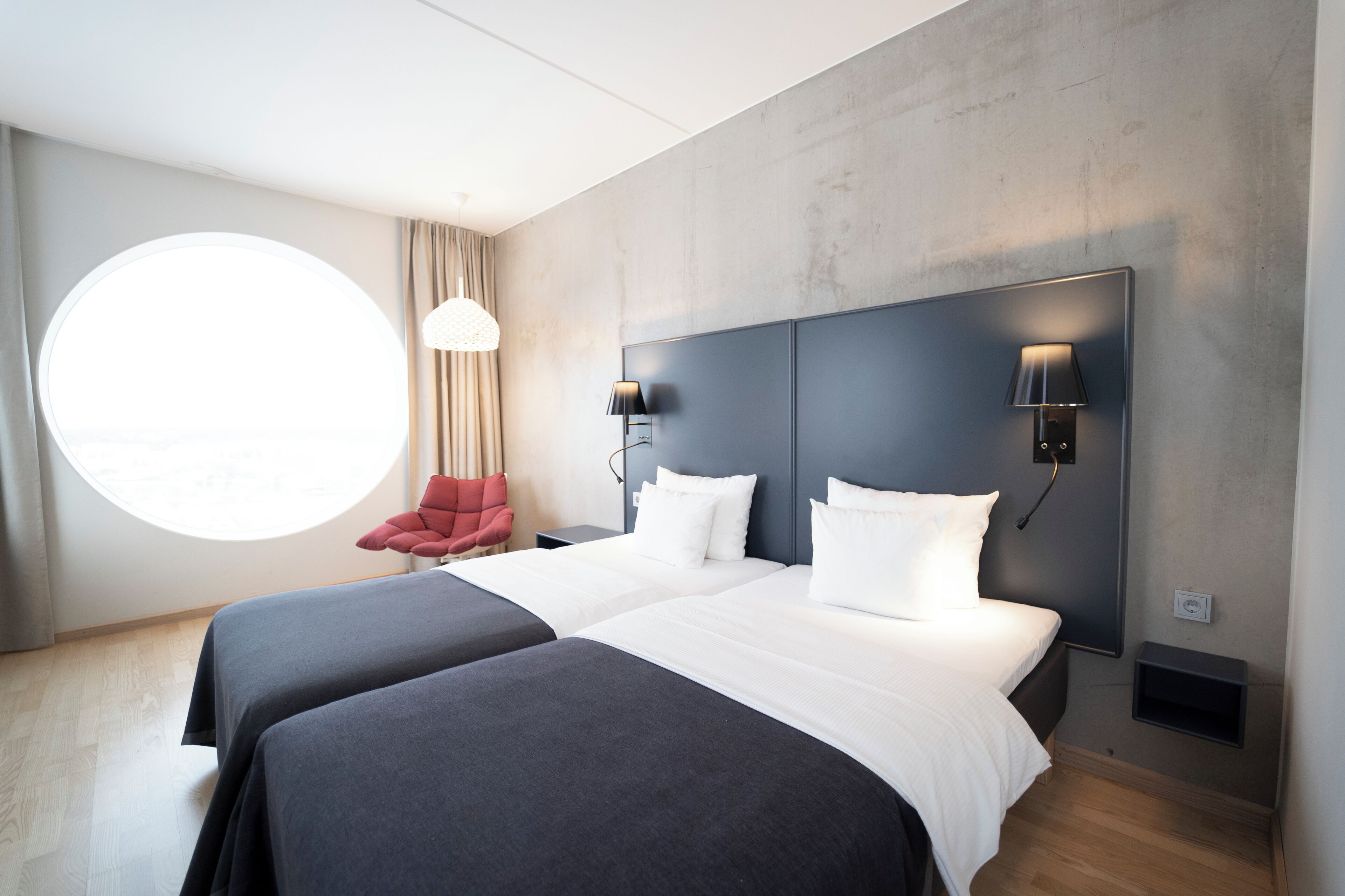A modern hotel room with two beds, a round window, a red chair, and a wall-mounted lamp.