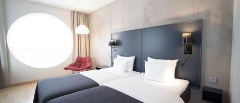 A modern hotel room with two beds, a round window, a red chair, and a wall-mounted lamp.
