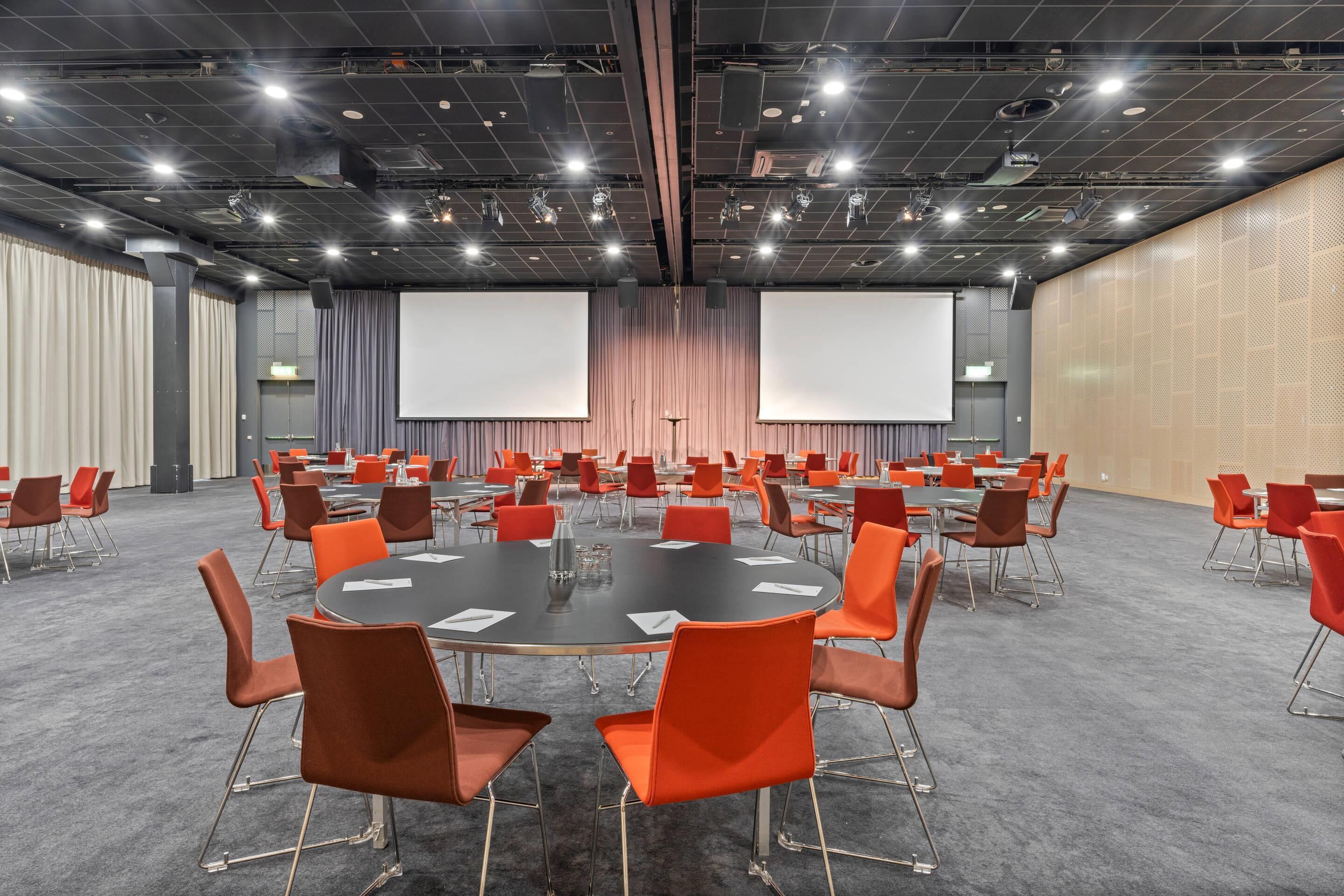 Un salón de conferencias con mesas redondas, sillas anaranjadas, una pantalla grande y un muro decorativo de madera.