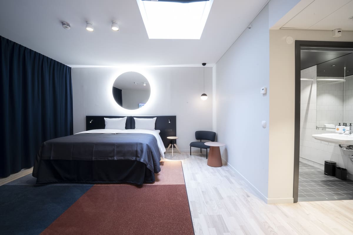 Ein modernes Hotelzimmer mit einem großen Bett, einem runden Spiegel, einer kleinen Sitzecke und einem Badezimmer mit Waschbecken und Spiegel.