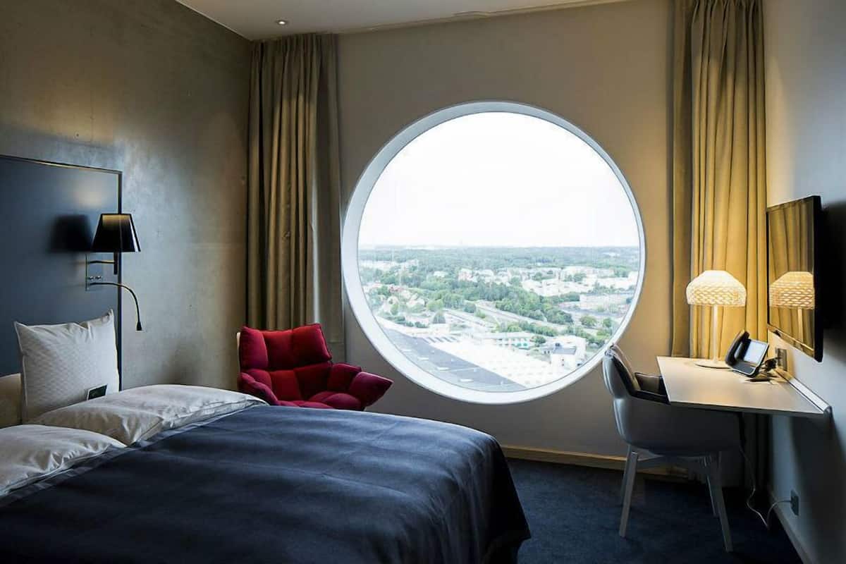 Ein Hotelzimmer mit Bett, Schreibtisch, Sessel und einem runden Fenster mit Blick auf die Stadt, sowie einer Lampe.
