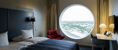 A hotel room with a bed, a desk, a chair, a round window with a city view, and a lamp.