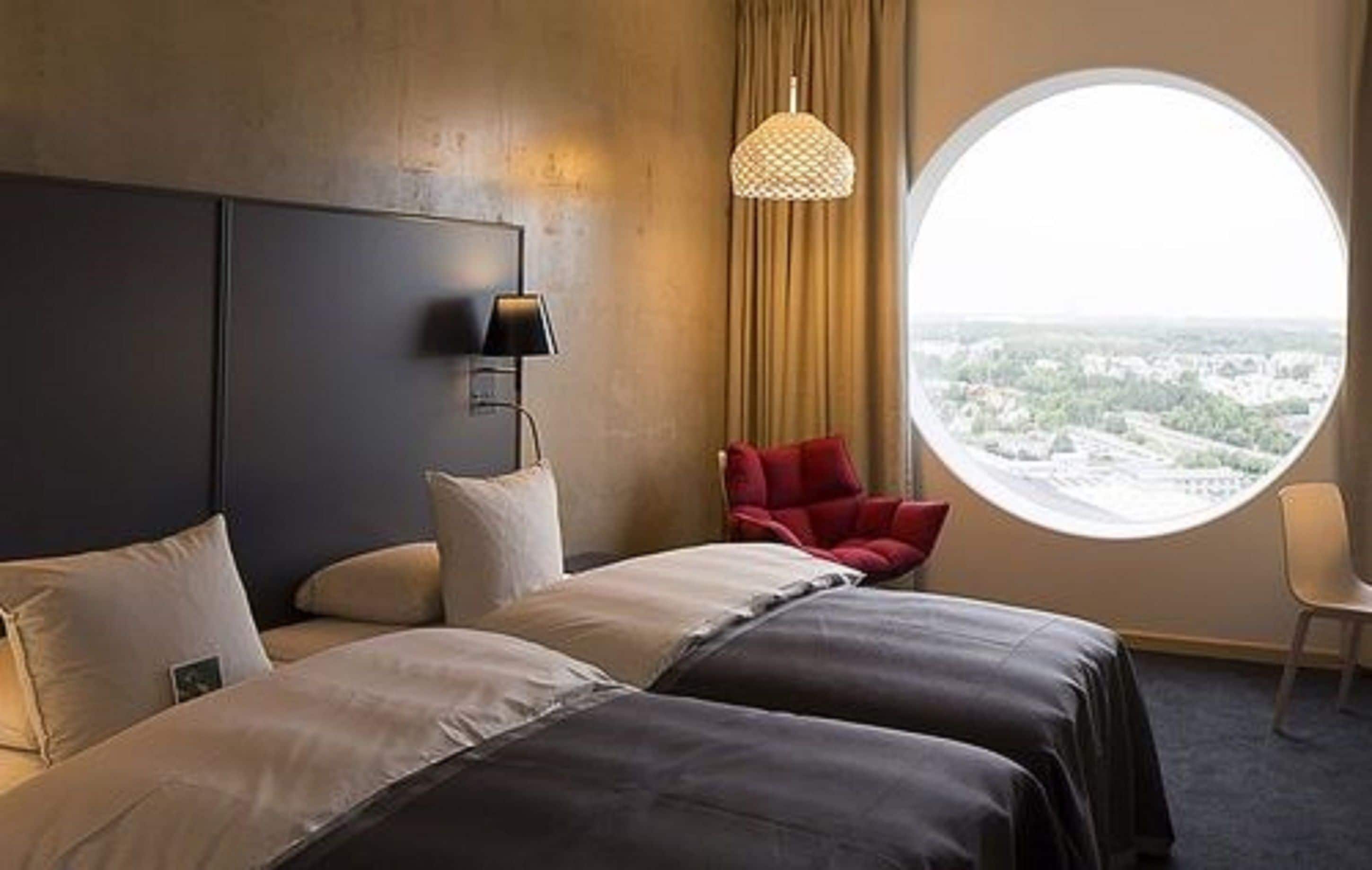 Een hotelkamer met een groot bed, een rode stoel, een rond raam en een wandlamp.