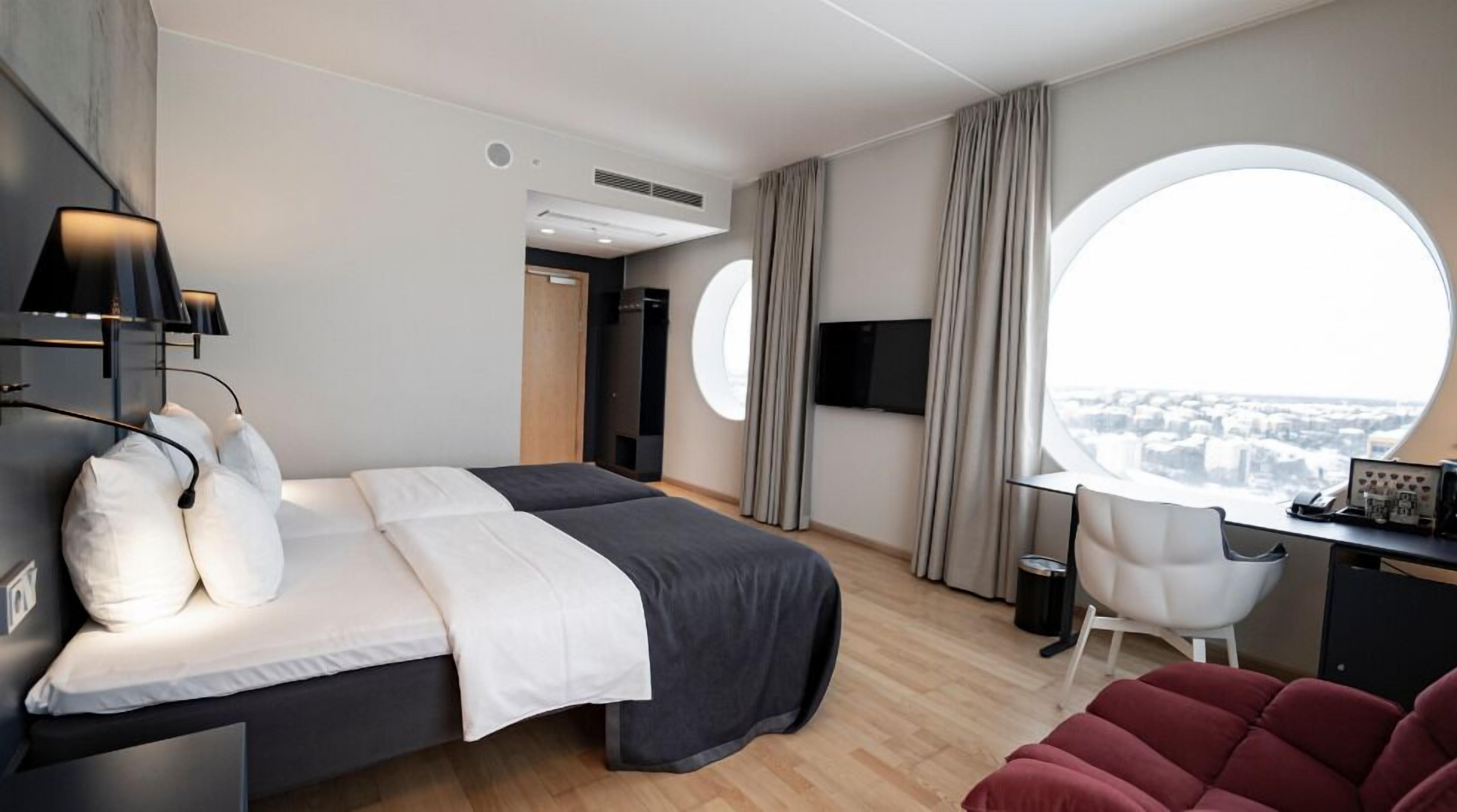 Une chambre d’hôtel moderne équipée d’un lit, d’un bureau, d’une chaise et d’une grande fenêtre donnant sur la ville.