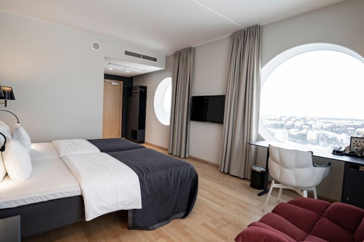 A modern hotel room with a bed, a desk, a chair, and a large window offering a city view.