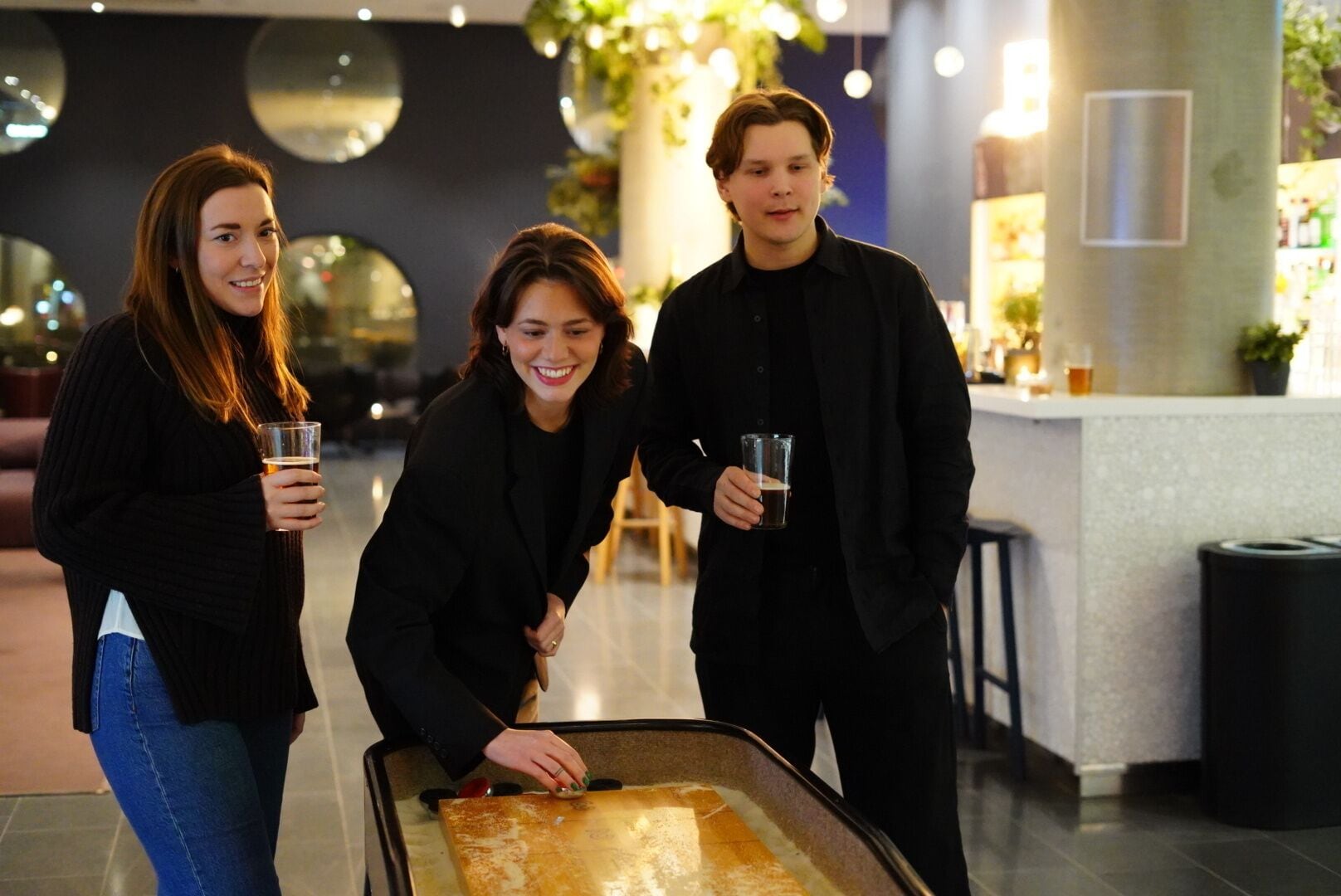 Drei Personen stehen um einen Tisch mit Getränken, im modernen Innenbereich mit einer Bar und Pflanzen im Hintergrund.