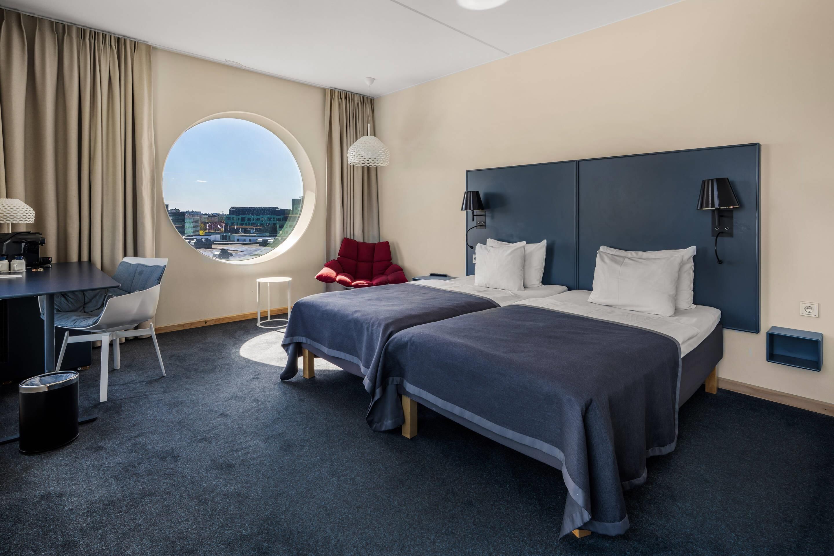 A hotel room with two beds, a desk, a chair, and a view of the city.