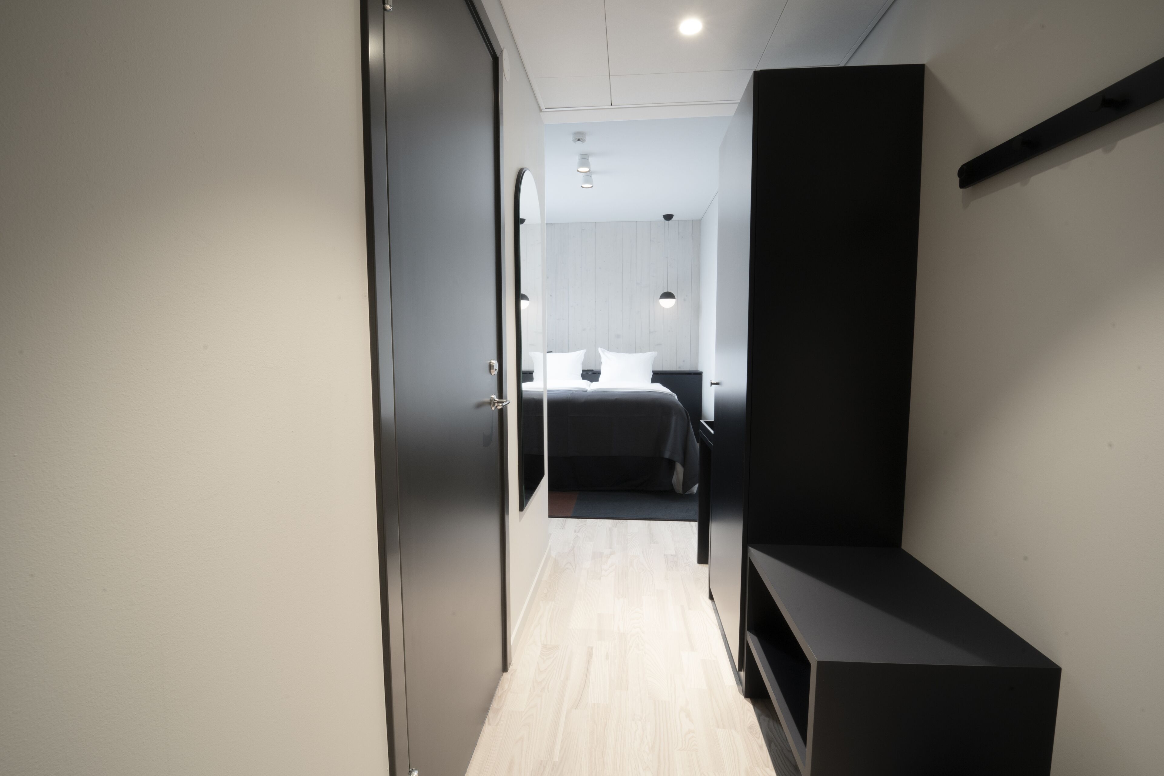 A modern hotel room with a minimalist design, featuring a bed with white linens, a dark console table, and a light-colored wooden floor.