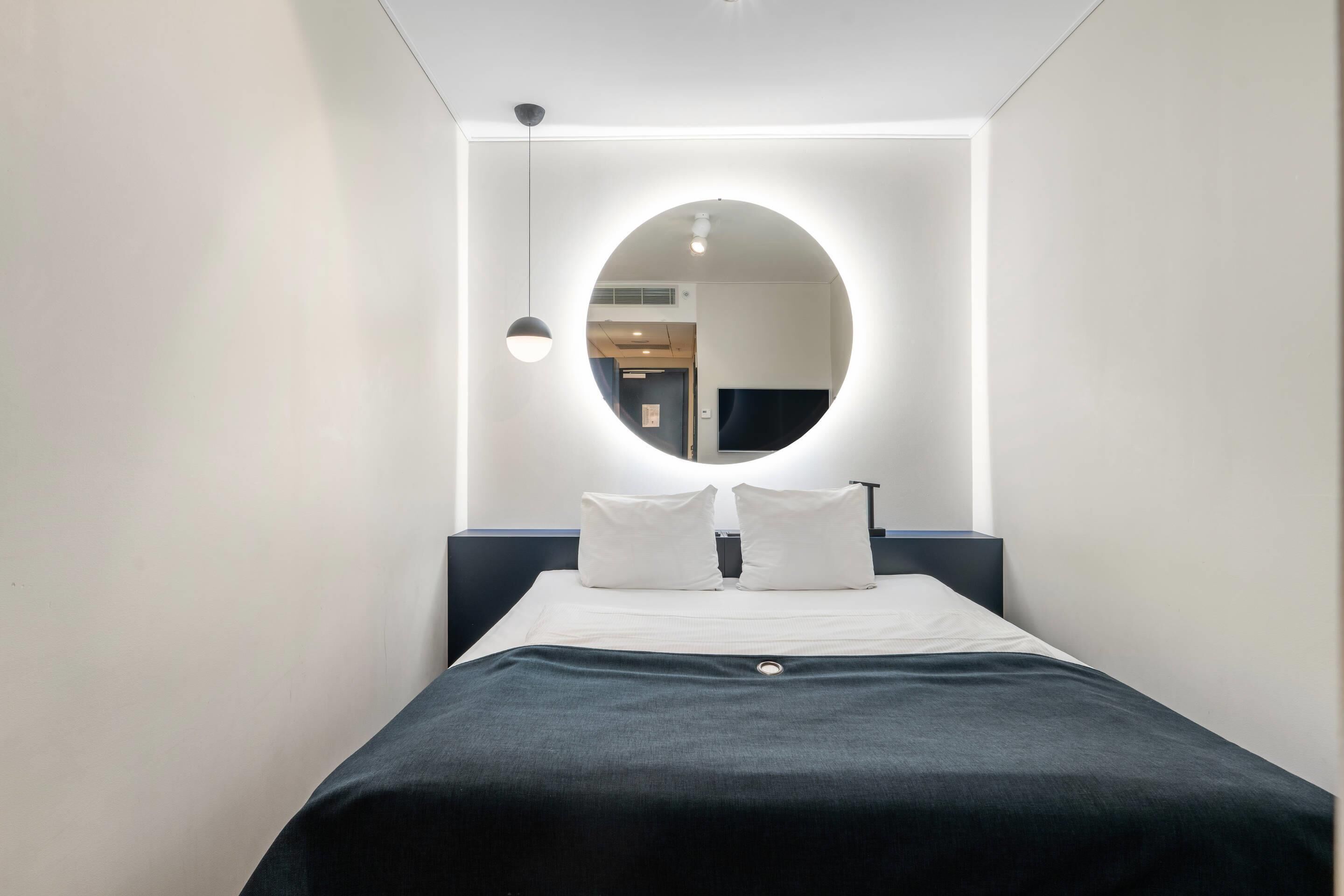 Hotellihuone, jossa on sänky, pyöreä peili ja riippuvalaisin.