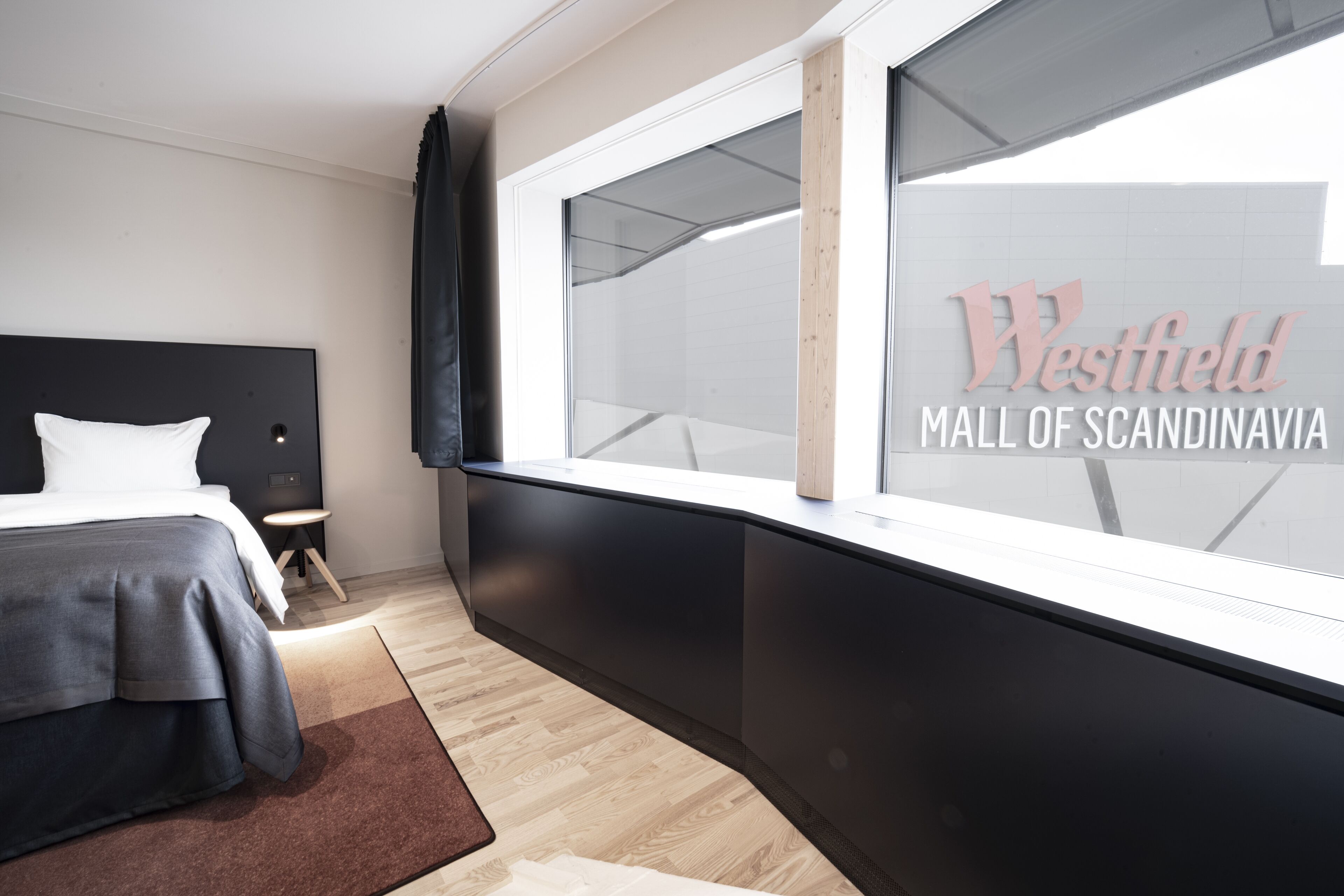Habitación de hotel moderna con una ventana grande, una cama, una mesita de noche y vistas al cartel del Westfield Mall of Scandinavia.