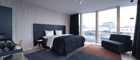 A modern hotel room with a large bed, a desk, and a sofa. The room has large windows with a city view.