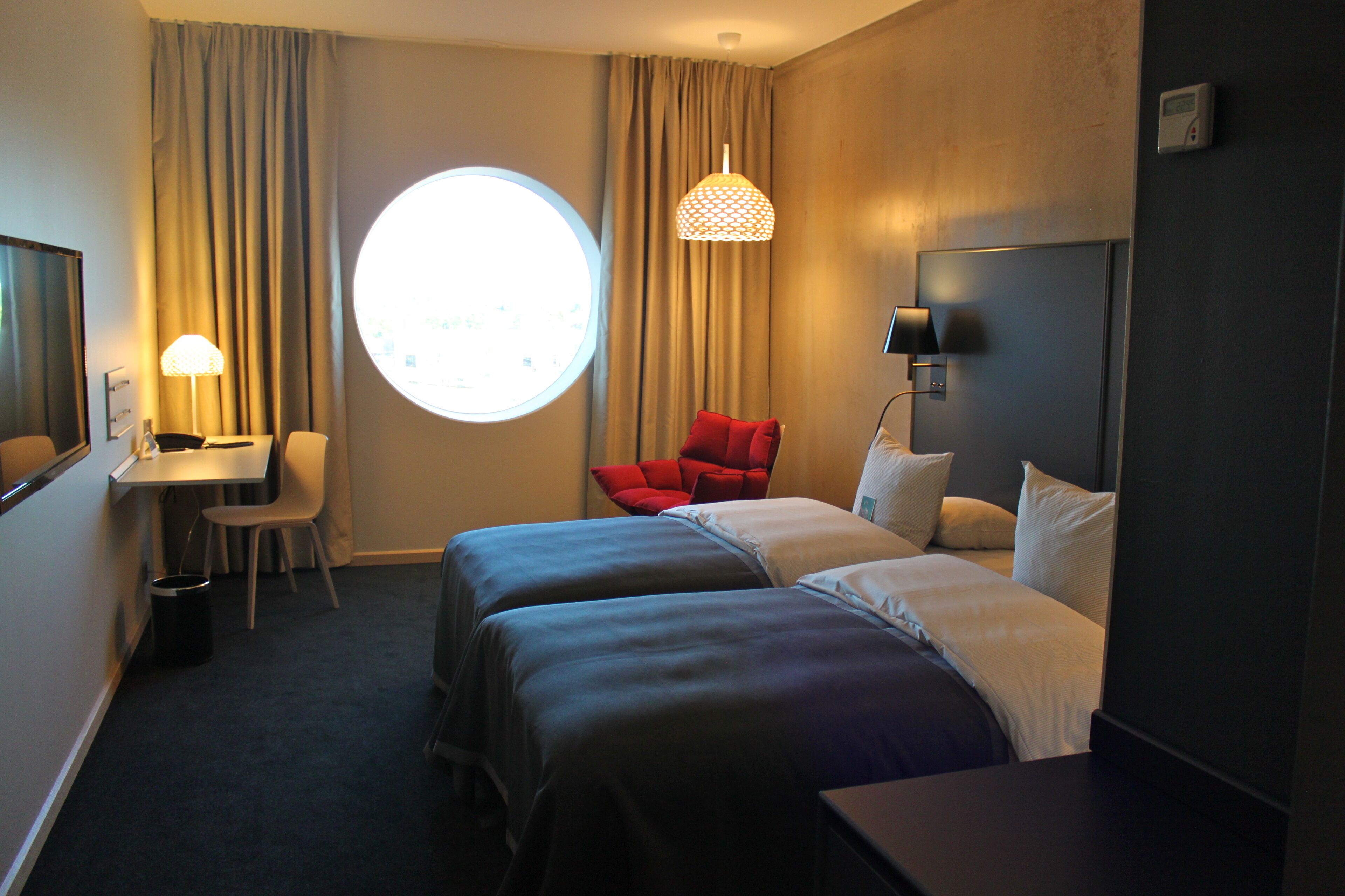 Habitación de hotel con dos camas, un escritorio, una silla, un ventanal redondo y una lámpara.