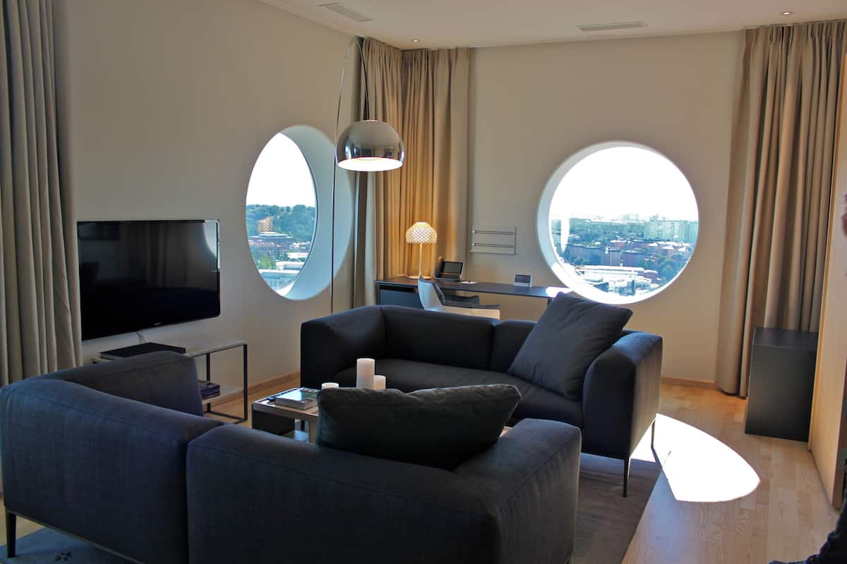Ein modernes Wohnzimmer mit einem Flachbildfernseher, zwei runden Fenstern mit Blick auf die Stadt, einem Sofa und einem Schreibtisch mit einem Laptop.