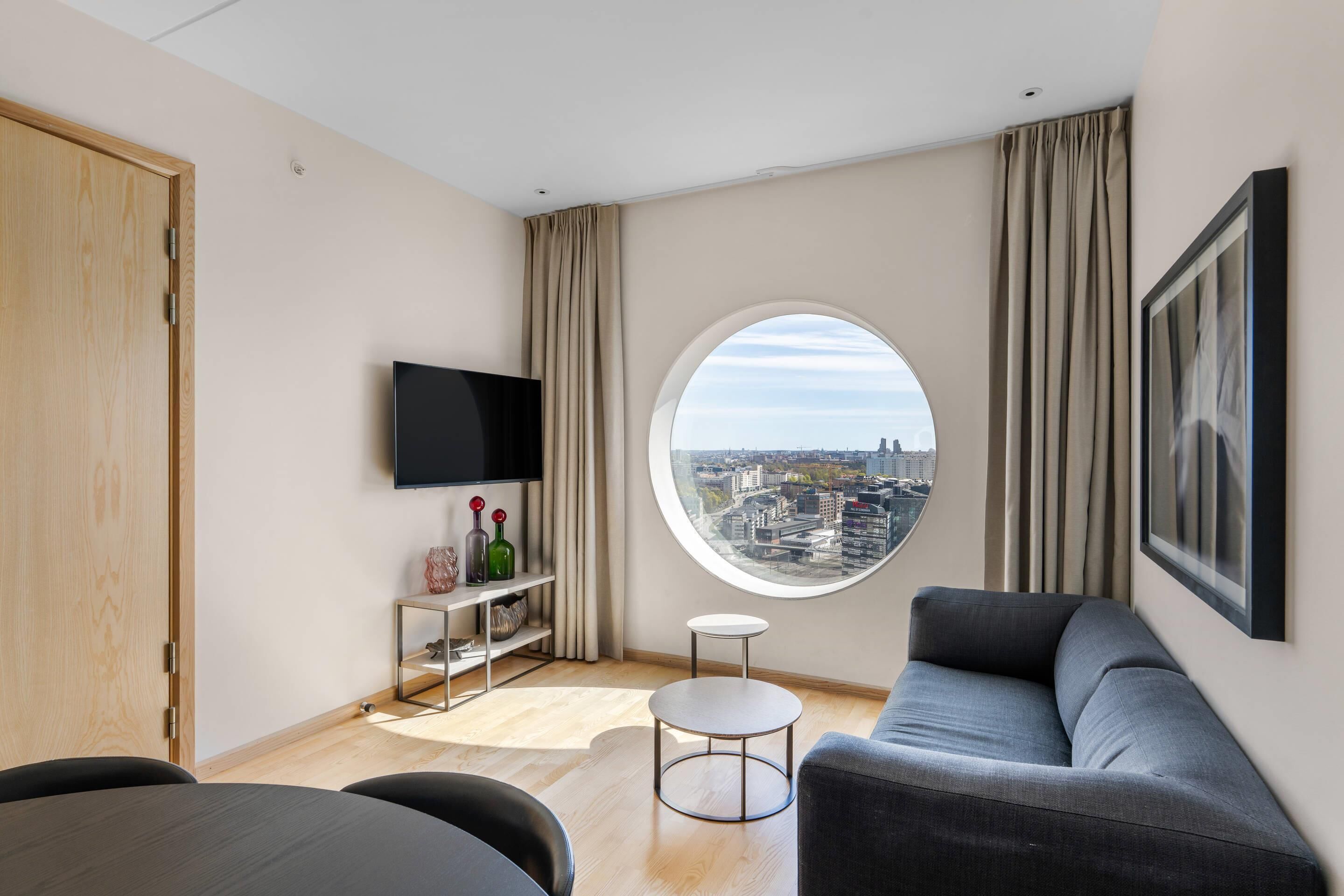 A modern living room with a city view, a flat-screen TV, a round window, and a grey sofa.