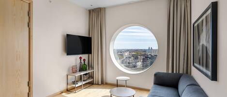 A modern living room with a city view, a flat-screen TV, a round window, and a grey sofa.