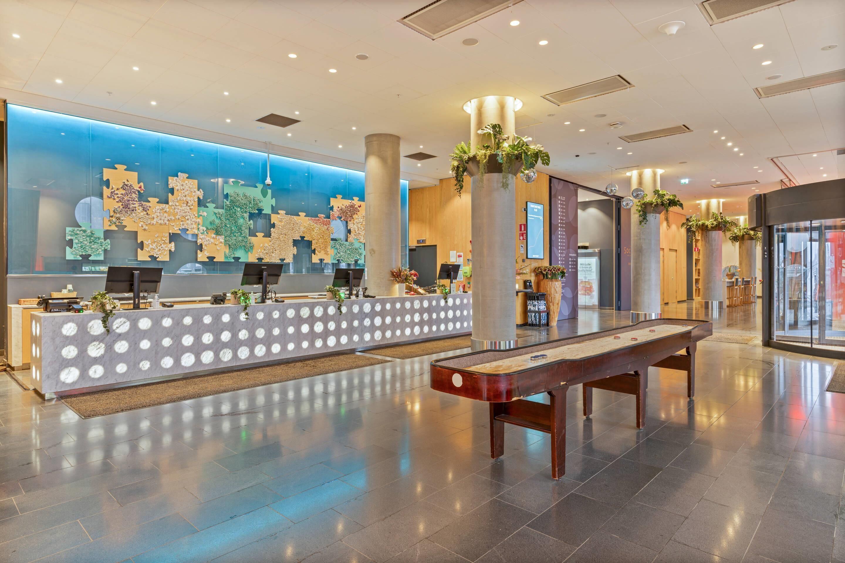 A hotel reception area with a modern design, featuring a reception desk, a digital information board, and a shuffleboard table.