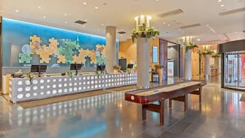 A hotel reception area with a modern design, featuring a reception desk, a digital information board, and a shuffleboard table.