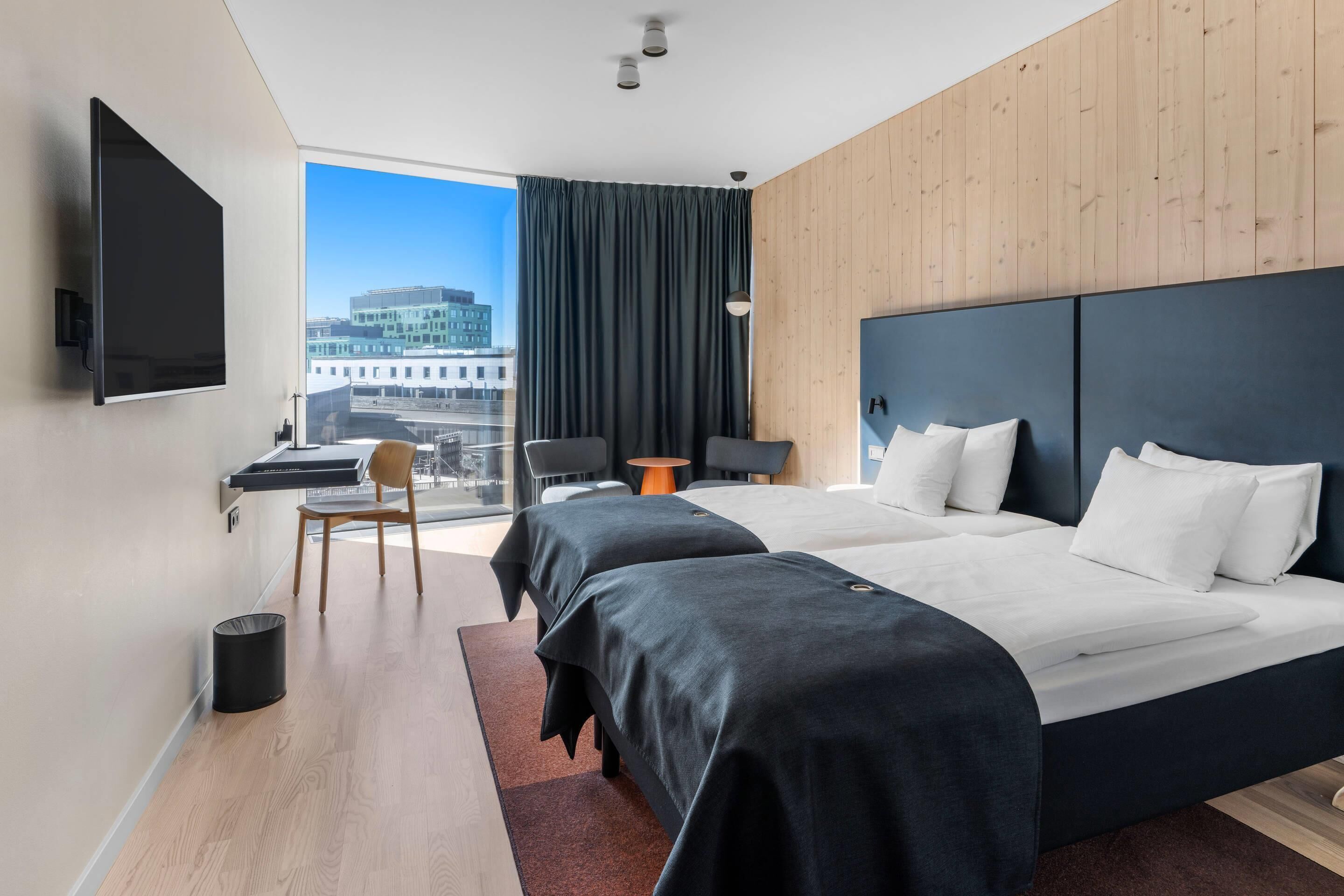Habitación de hotel con dos camas, un escritorio, una silla, televisión y vista a la ciudad.