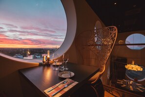A dining table with a view of the sunset through a large circular window, a chair with a decorative backrest, and a glass table with a candle. - Quality Hotel Strawberry Arena (Solna)