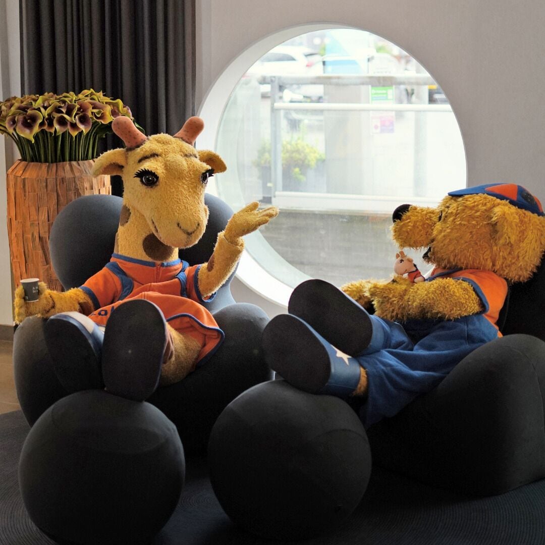 Två mjuka gosedjur sitter på svarta sittpuffar i ett rum med ett stort fönster och en vas med blommor.